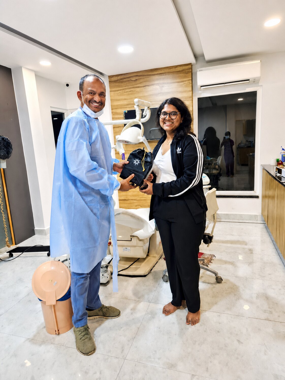 Prof. Dr S. Srinivas, gowned, handing a patient her aligner pouch beside the chair in a treatment room.