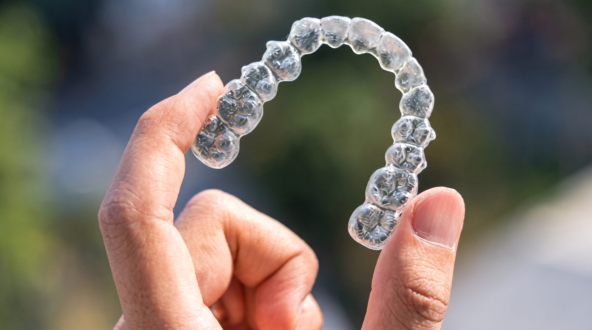 A clear removable aligner tray held up to the light between finger and thumb, the individual tooth sockets visible along its arch.