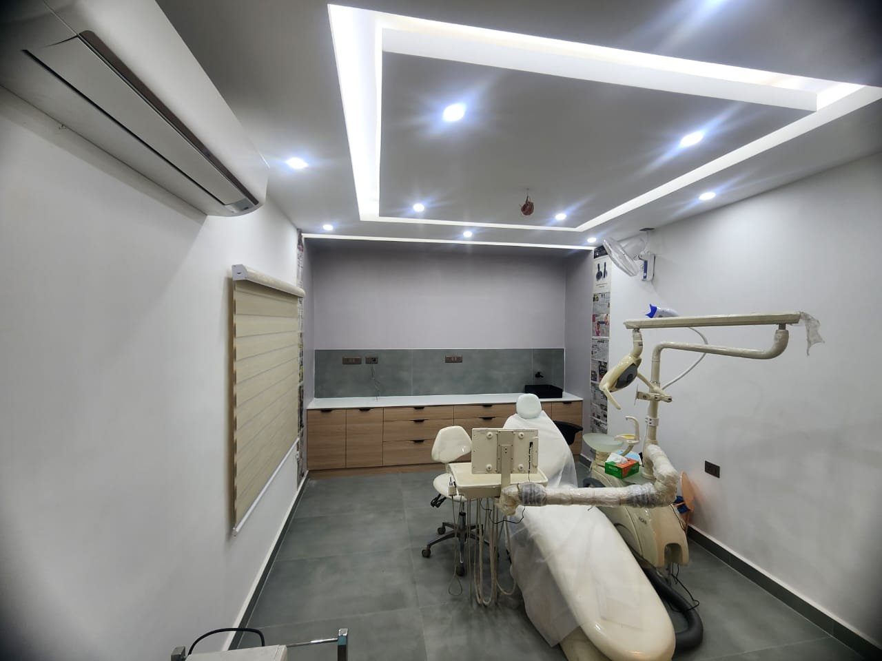 A treatment room with a cove-lit ceiling, timber cabinetry, a roller blind and a white dental chair.