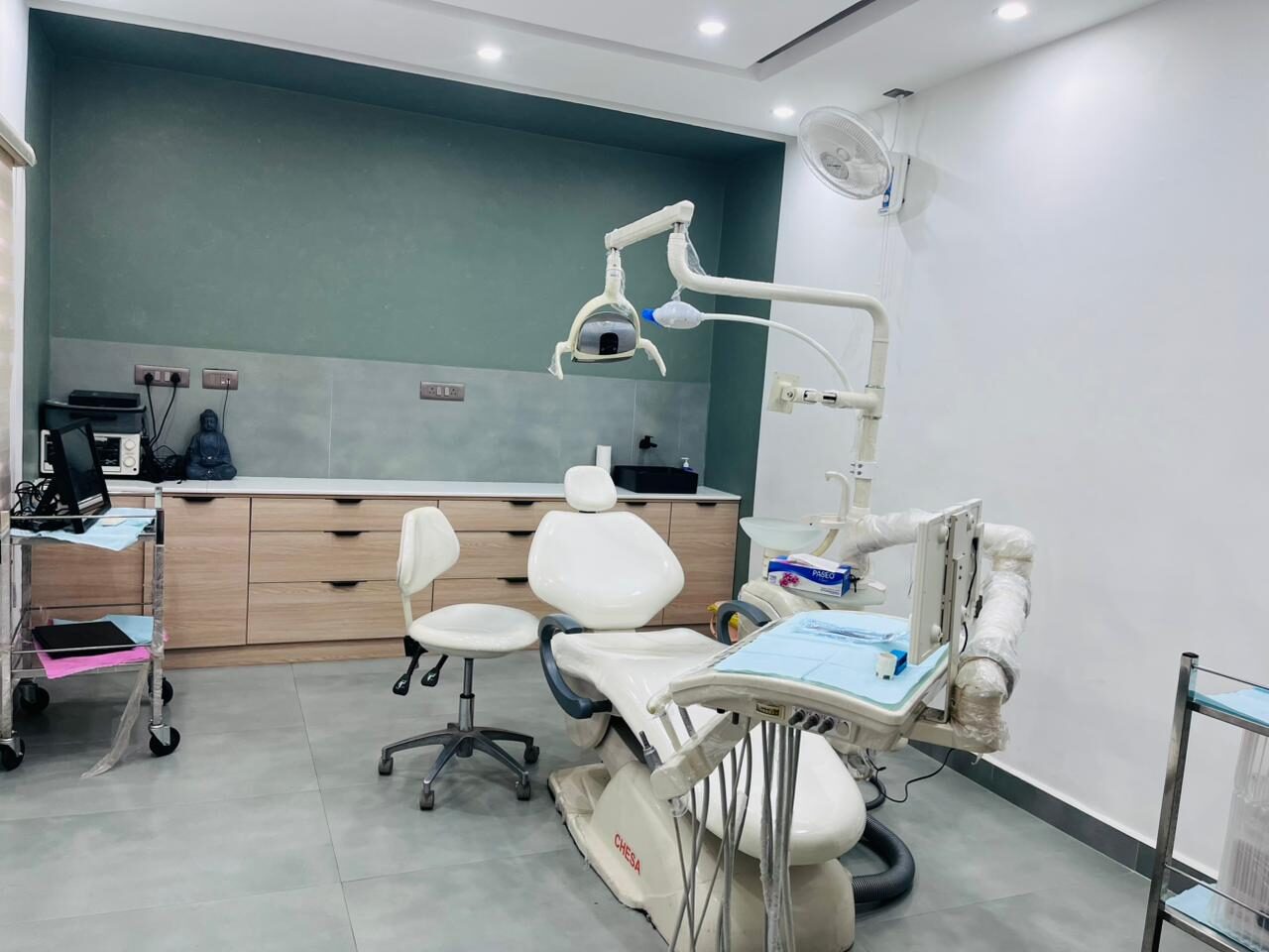 A treatment room with a deep teal wall above a tiled splashback, timber cabinetry running along it, a cream dental chair and operator’s stool under the operating light, and a wall fan above.
