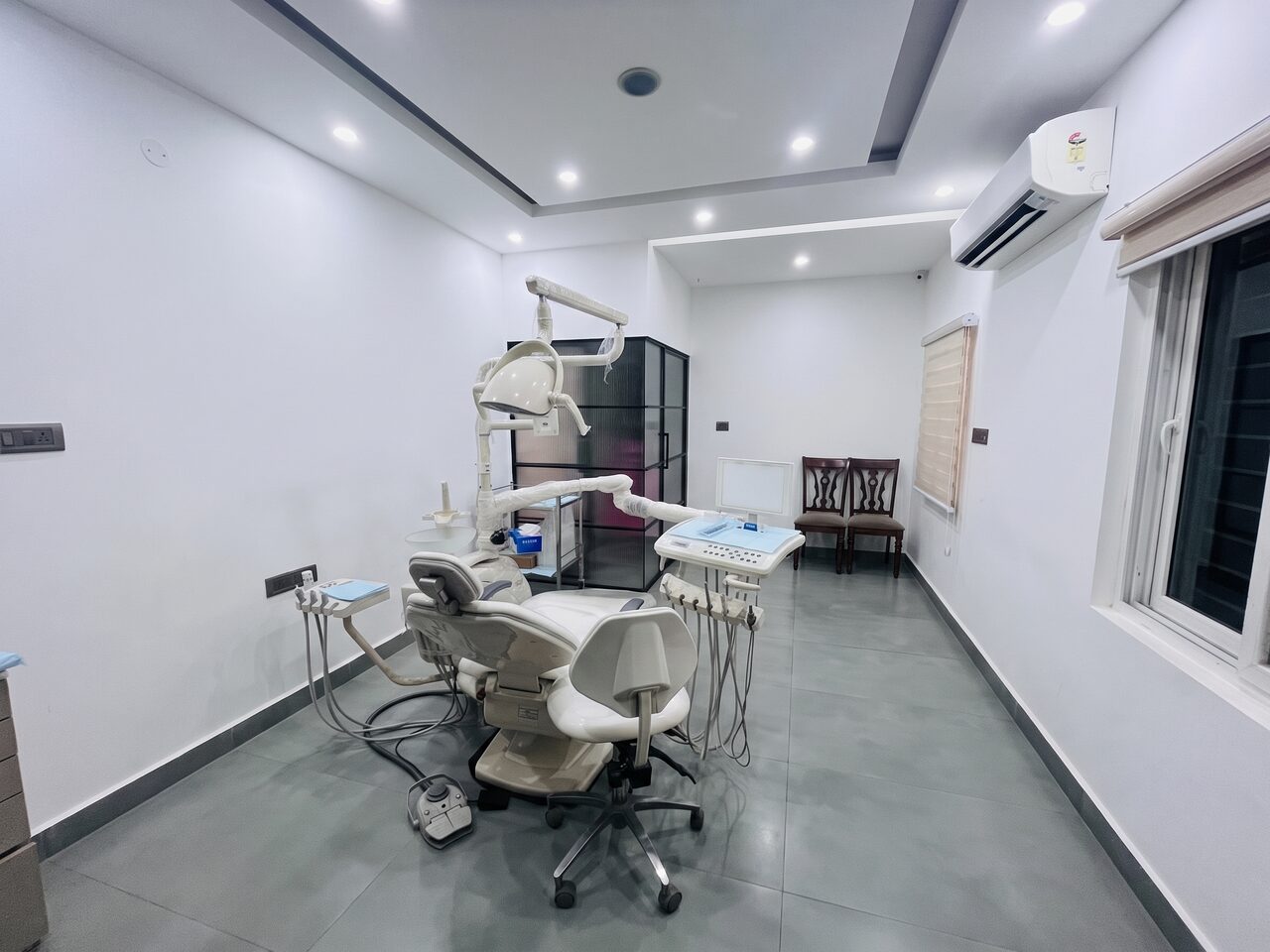 A treatment room looking down its length: the dental chair under its operating light, a black-framed glazed partition behind it and two wooden chairs against the far wall.