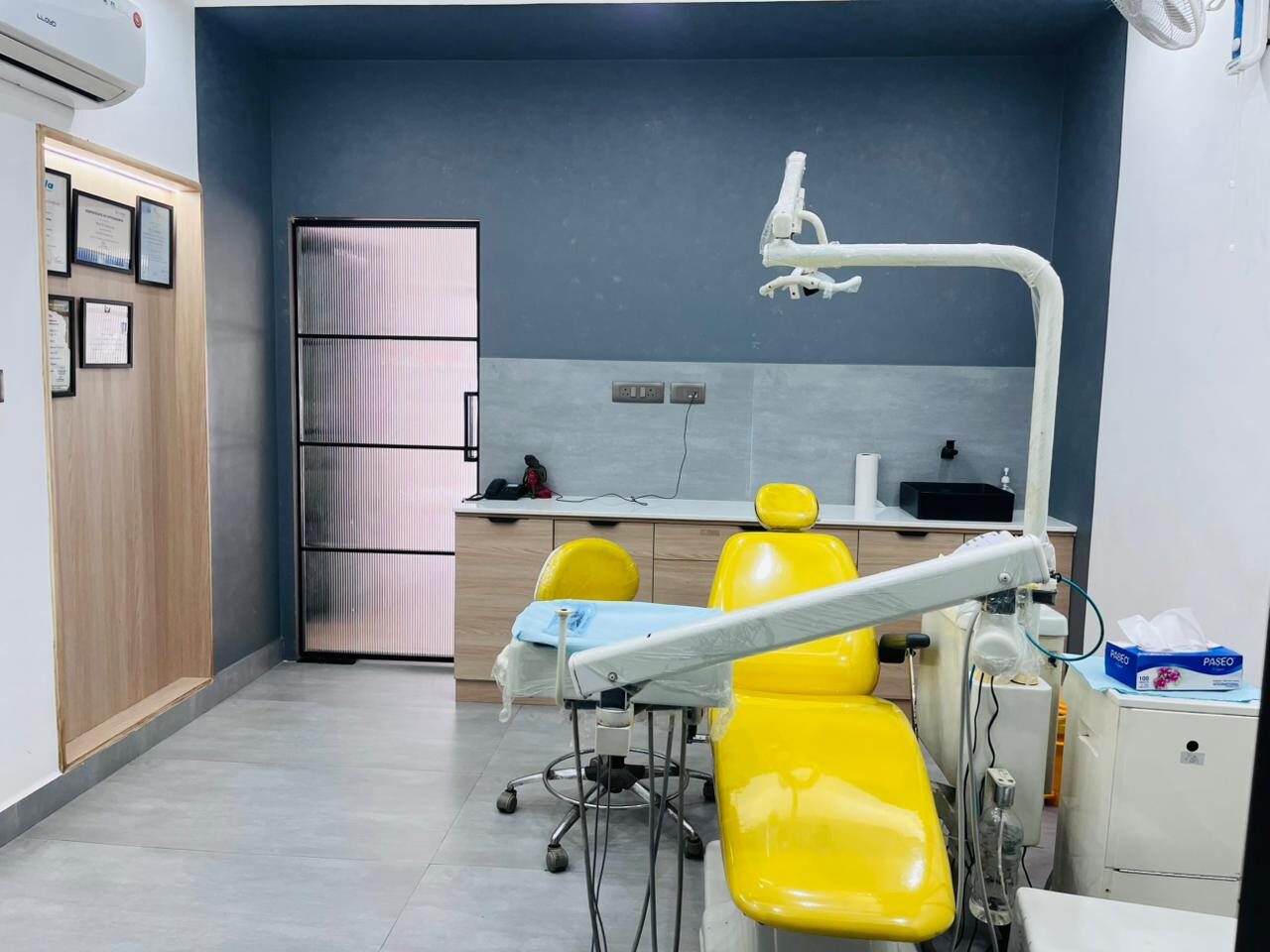 A treatment room with a slate blue wall, a pink ribbed-glass door in a black frame, timber cabinetry along the splashback and a bright yellow dental chair.