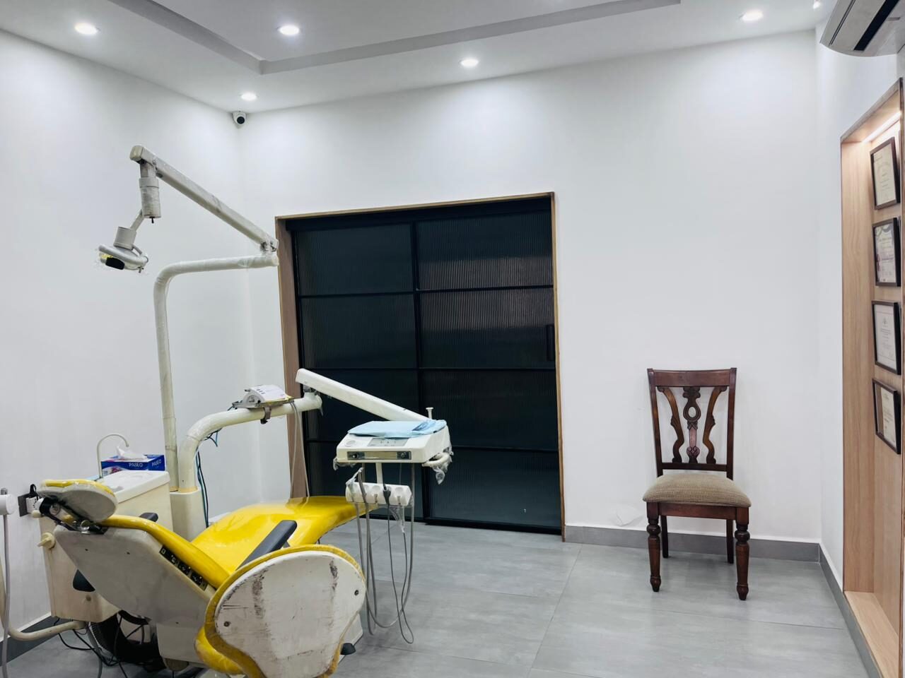 A treatment room with a yellow dental chair and its instrument unit, a black-framed ribbed-glass door in a timber surround, and framed certificates on the wall beside a wooden chair.
