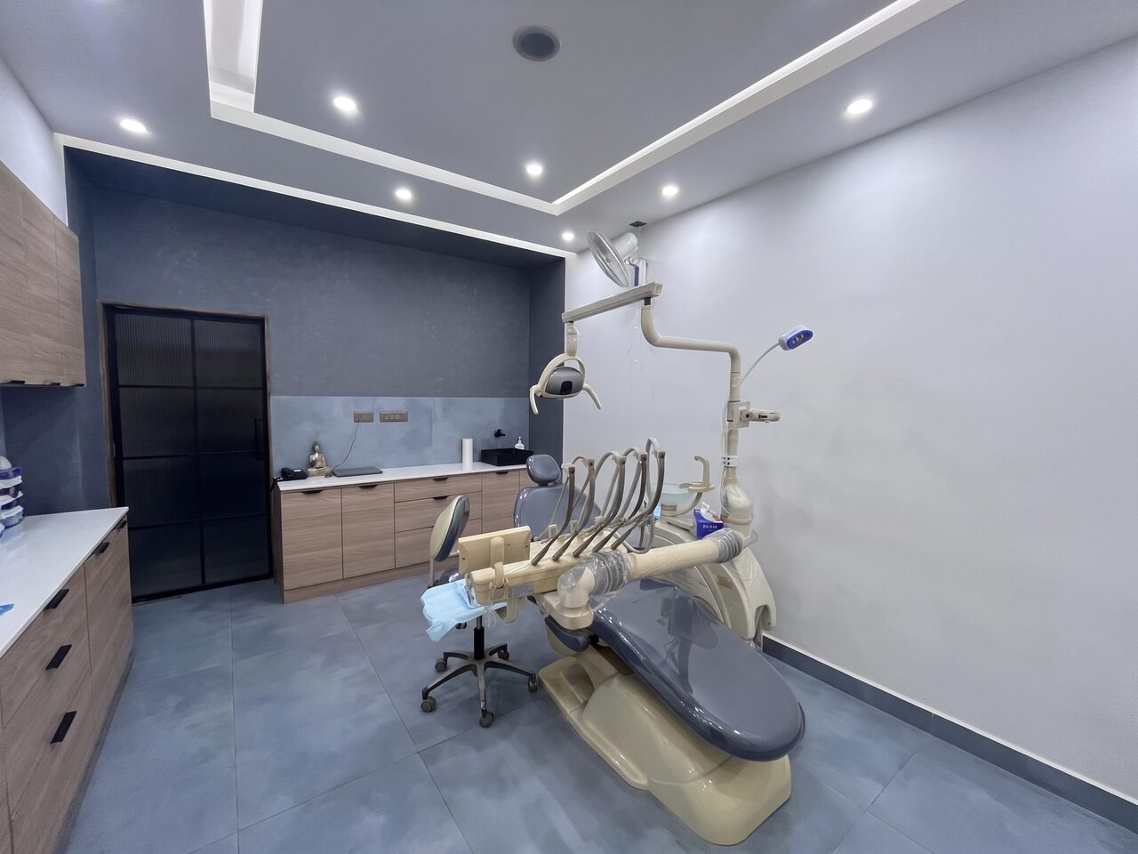 A treatment room with a dark slate wall above a pale blue splashback, timber cabinetry, and a cream and grey dental chair under its operating light.