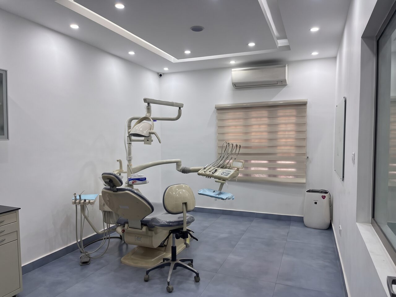 A treatment room with a roller blind drawn across the window, an air conditioner above it, and a cream and grey dental chair with its instrument tray.