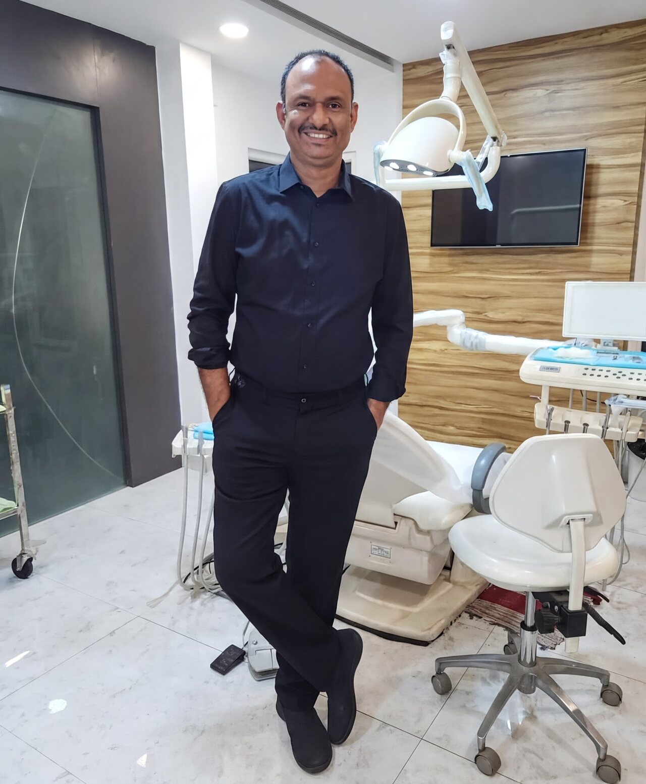 Prof. Dr S. Srinivas standing beside the chair in his operatory at Align Dental Care, with the operating light and instrument tray behind him.