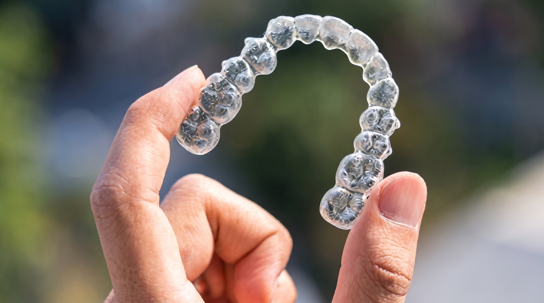 A clear removable aligner tray held up to the light between finger and thumb, the individual tooth sockets visible along its arch.